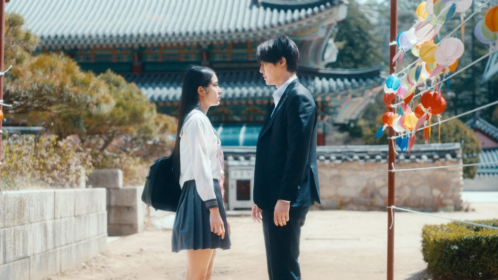 伝統的な寺院を背景に、制服を着た若い女性とスーツ姿の男性が向き合って話しています。