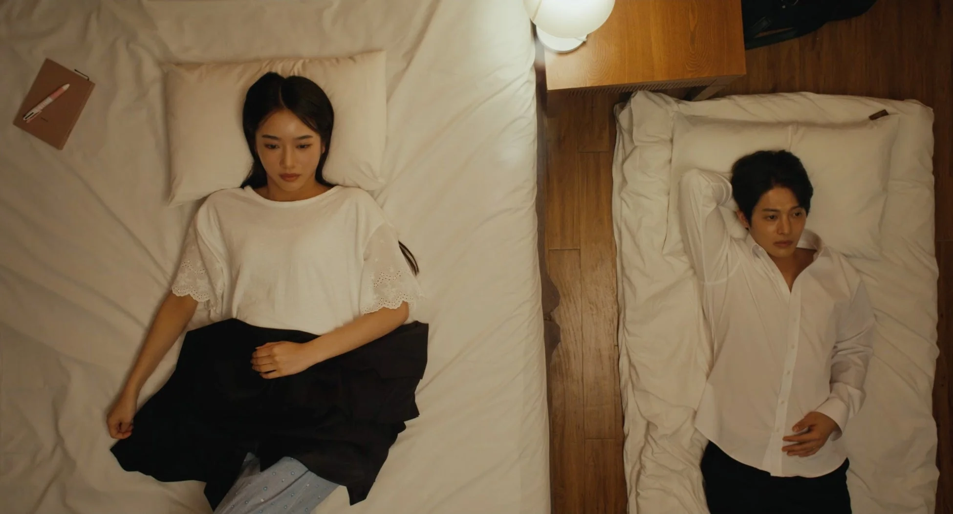 寝室で男女が別々のベッドに横たわり、それぞれ物思いにふけっている様子。