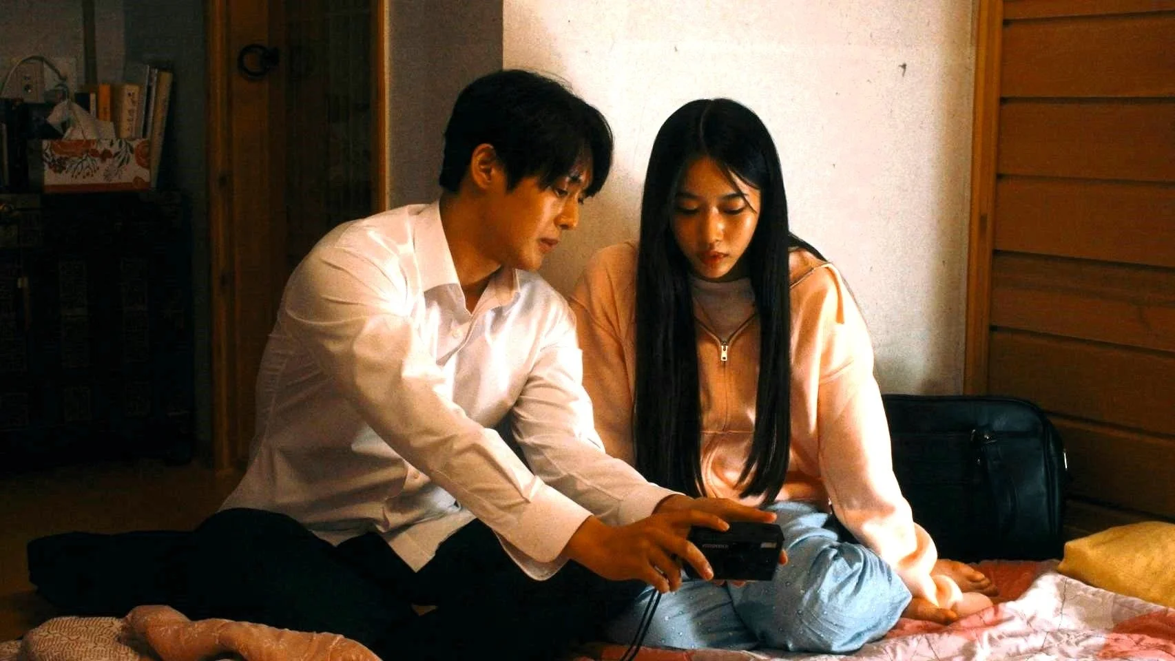 若い男女が部屋の床に座り、男性が持つ小さな黒いデバイスを一緒に見ている様子です。