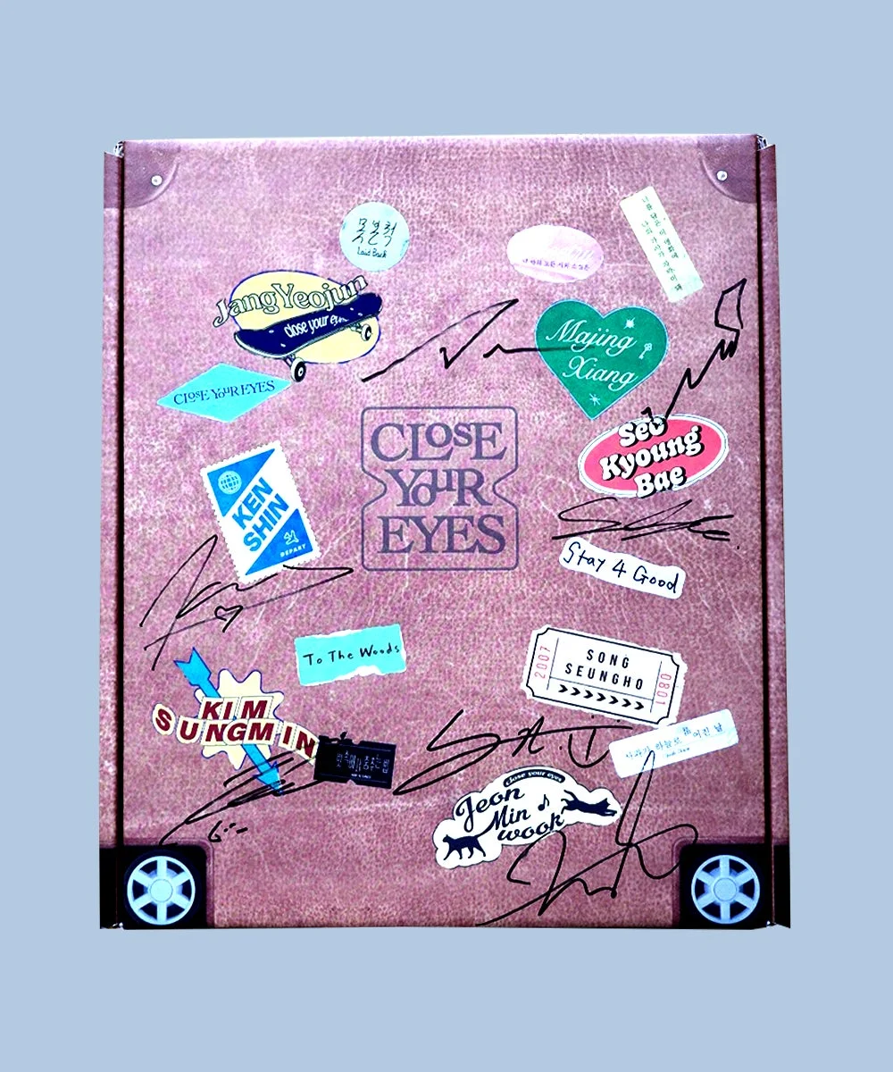 ピンクのスーツケースのような箱型オブジェクトに、多数のカラフルなステッカーと複数のサインが施された画像です。中央には「CLOSE YOUR EYES」という文字があり、全体的に旅行の記念品やファンアイテムのようなパーソナルな雰囲気を醸し出しています。
