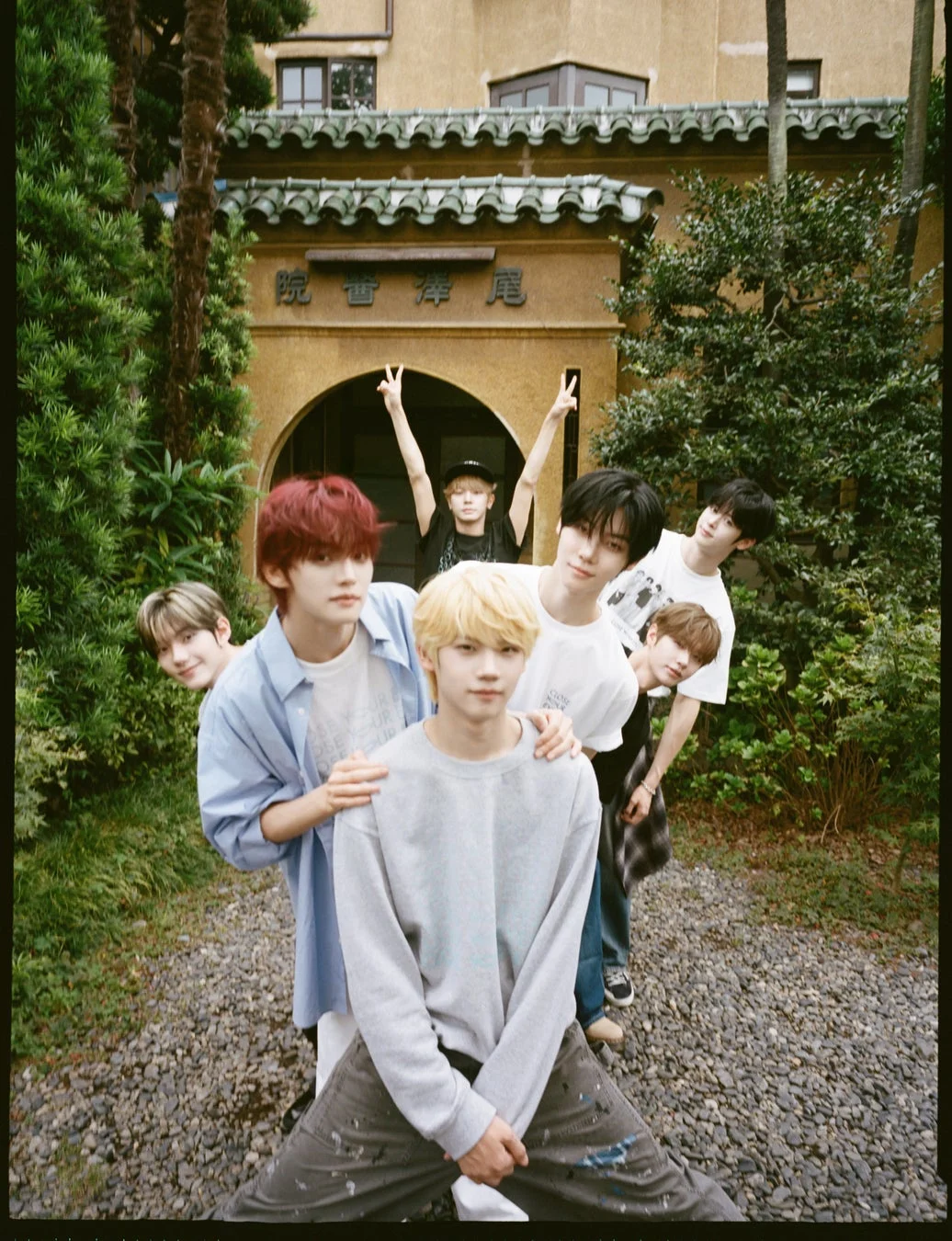 複数の若い男性が伝統的な建築物の前で楽しそうにポーズをとっています。カジュアルな服装で、ピースサインをするメンバーもおり、和やかな雰囲気が伝わります。