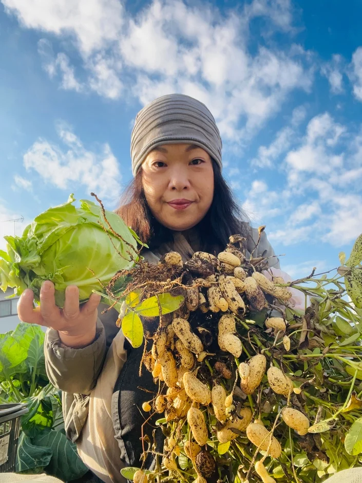 青空の下、女性が収穫したばかりのピーナッツとキャベツを手に笑顔を見せている