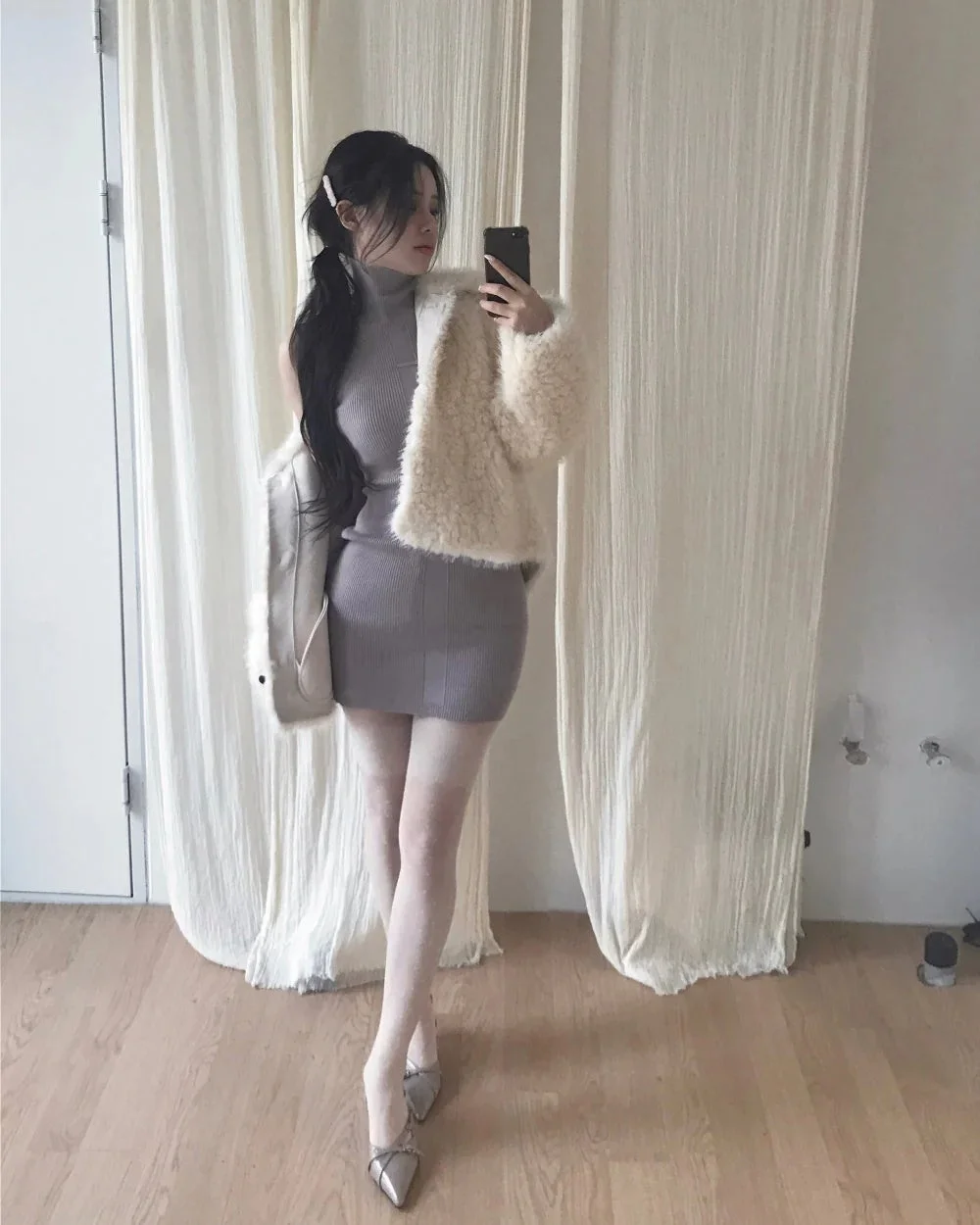 グレーのタイトなタートルネックミニワンピースと白いふわふわのジャケットを着用した女性