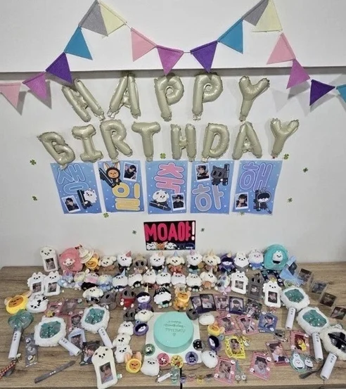 K-POPアイドルの誕生日を祝う飾り付けがされた部屋で、沢山のファンアイテムが並べられたテーブルが写っています