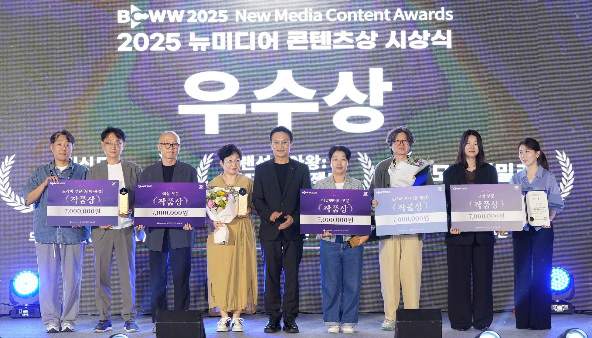 2025年ニューメディアコンテンツアワードの授賞式で、優秀賞を受賞した関係者たちがステージに上がり、賞状や700万ウォンの賞金パネル、花束を手に記念撮影をしている集合写真
