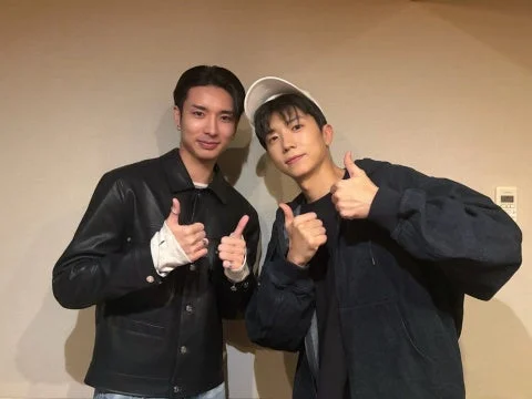 壁を背景に、2人の男性が笑顔でカメラに向かってサムズアップのポーズをとっています。カジュアルな服装で、楽しげな雰囲気が伝わります。