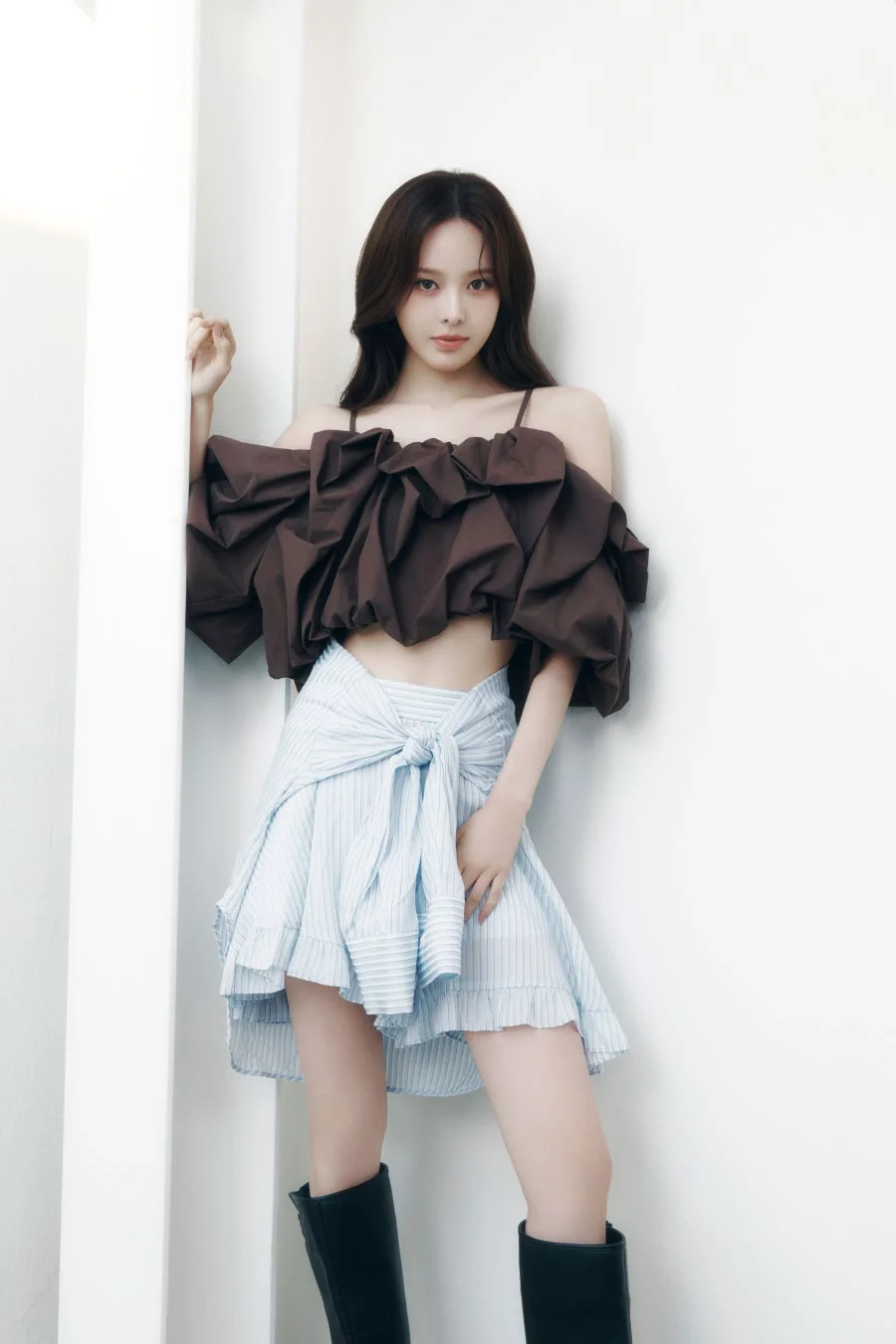 白い壁を背景にポーズをとる若い女性の全身ポートレート。ブラウンのフリル付きオフショルダークロップトップと水色のストライプ柄ミニスカート、黒いロングブーツを着用しており、スタイリッシュなファッションが特徴的です。