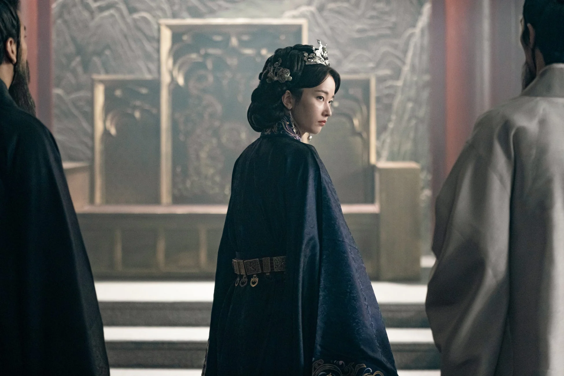 豪華な伝統衣装の女性が男性二人に囲まれ振り返る場面