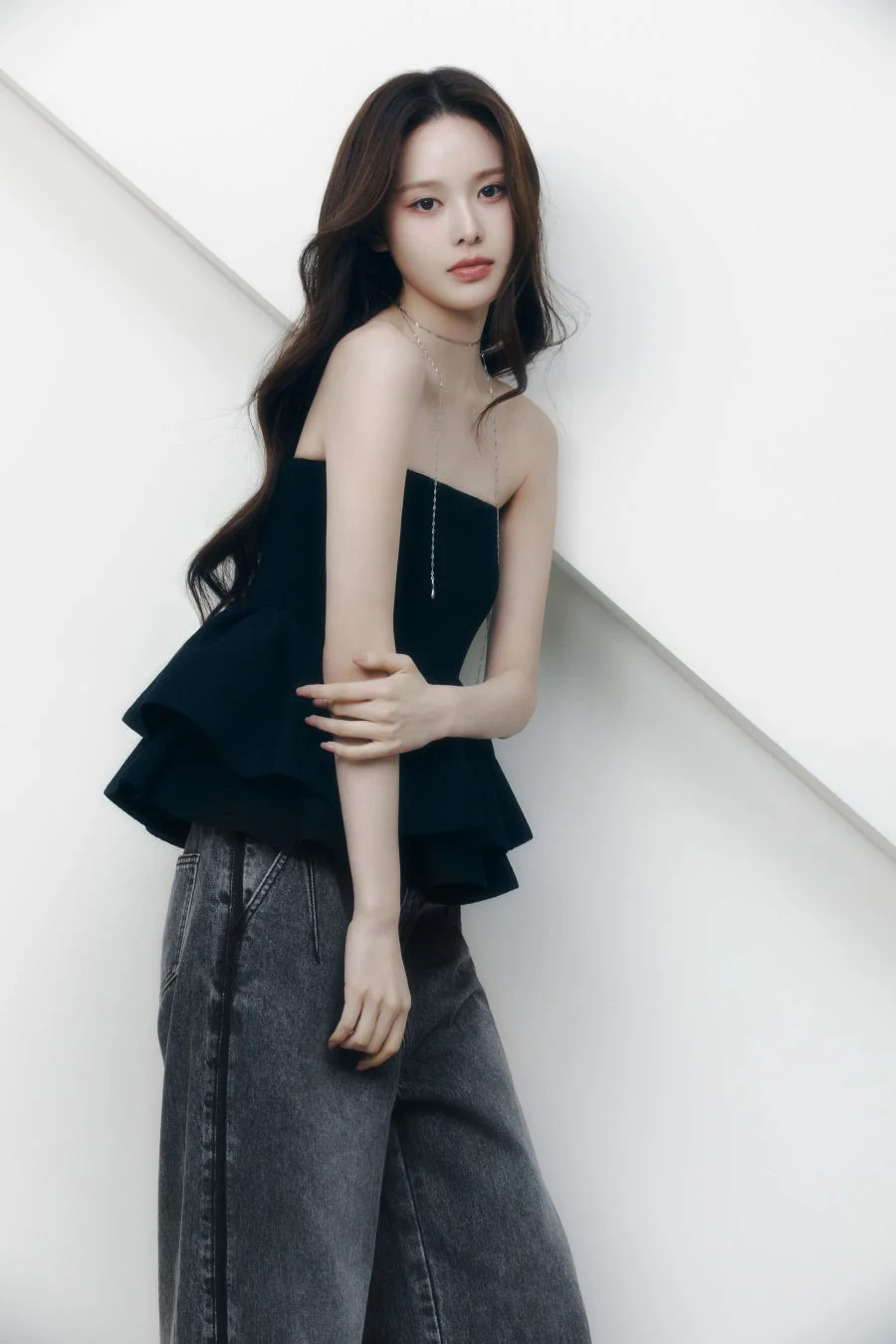 白い幾何学的な背景の前でポーズをとる、黒のストラップレスペプラムトップスとグレーのワイドレッグジーンズを着用した若い女性の全身ポートレートです。