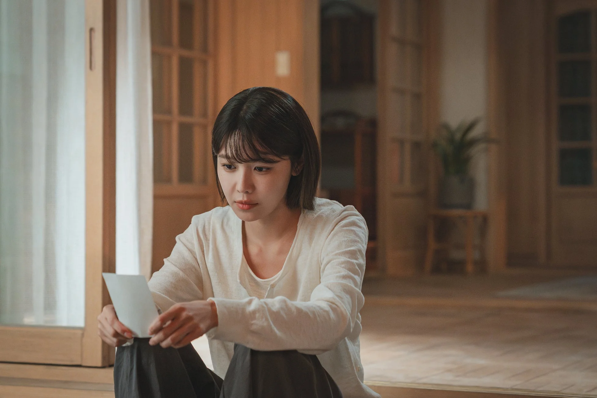 和室のような空間で白い紙片を手に考え込む若い女性