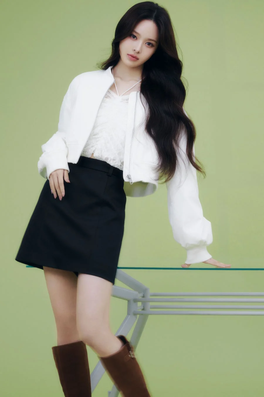 緑色の背景をバックに、白いボンバージャケットと黒いミニスカートを身につけた若い女性が立っています。長い黒髪が印象的で、ガラスのテーブルに軽く手を置くポーズをとっています。