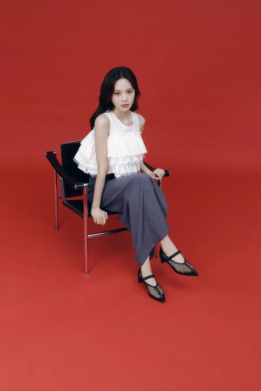 赤い背景のスタジオで、黒い椅子に座る女性の全身ポートレートです。白いフリルのトップスとグレーのワイドパンツ、黒いメッシュのパンプスを着用しており、ファッション性の高い一枚です。