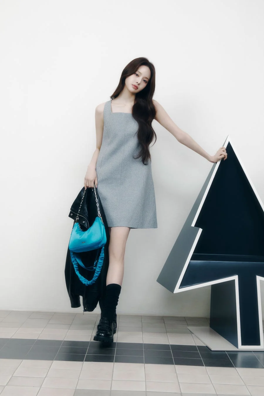 白い壁の前でポーズをとる女性の全身像です。グレーのノースリーブミニドレスに黒いブーツを合わせ、ターコイズブルーのハンドバッグと黒いジャケットを持っています。