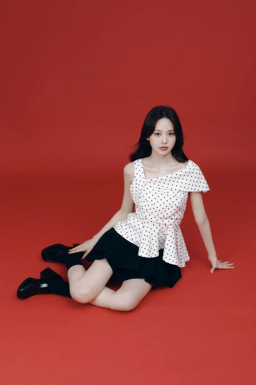 赤い背景のスタジオで、白地に黒のドット柄トップスと黒いスカートを着用した若い女性が座っています。カメラを見つめるポーズが印象的です。