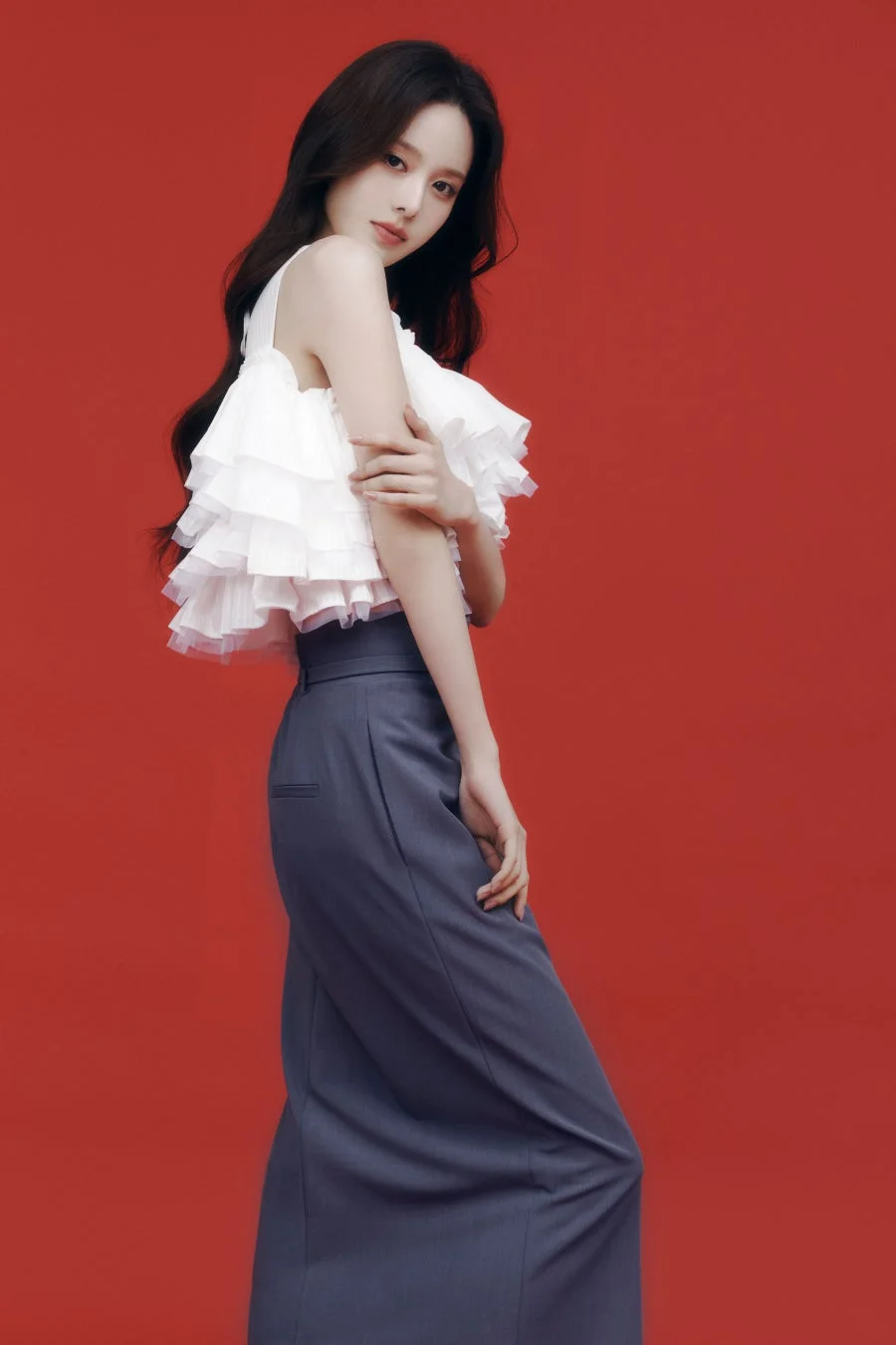 赤い背景の前でポーズをとる若い女性の全身ポートレート。白いフリルのトップスとダークグレーのワイドパンツを着用し、カメラに視線を向けています。