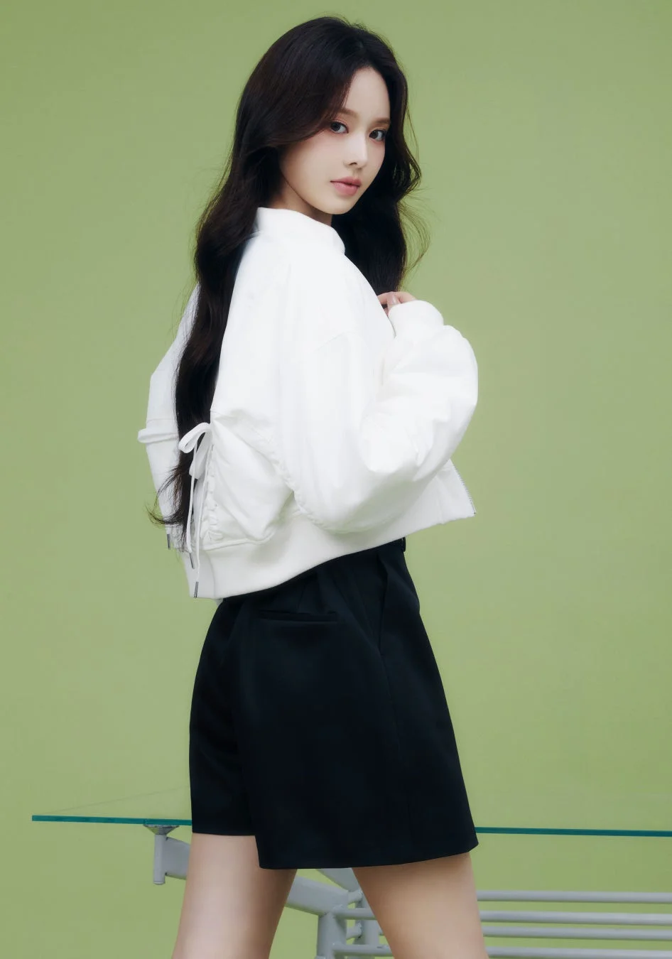 緑の背景を背に、白いクロップド丈のジャケットと黒いショートパンツを着用した若い女性が、カメラに視線を送っているポートレート。