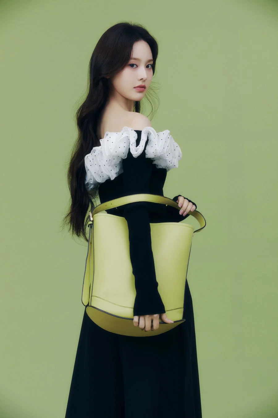 ライムグリーンのバッグを持った女性が、緑色の背景の前でポーズをとっています。黒いオフショルダートップに白いフリルとドット柄の装飾が特徴的で、エレガントな雰囲気を醸し出しています。