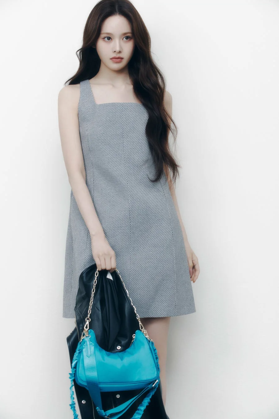 白い背景の前でポーズをとる若い女性の全身像です。彼女はグレーの柄入りノースリーブドレスを着用し、黒いレザージャケットと鮮やかな青いショルダーバッグを持っています。