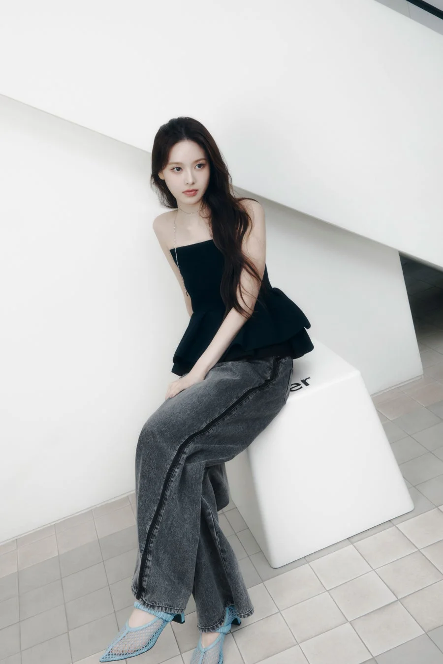 白い空間で、黒いチューブトップとダークグレーのワイドレッグジーンズを着用した若い女性が、白い台座に座っています。ライトブルーのメッシュシューズが特徴的で、全体的にモダンでミニマルな雰囲気のポートレートです。