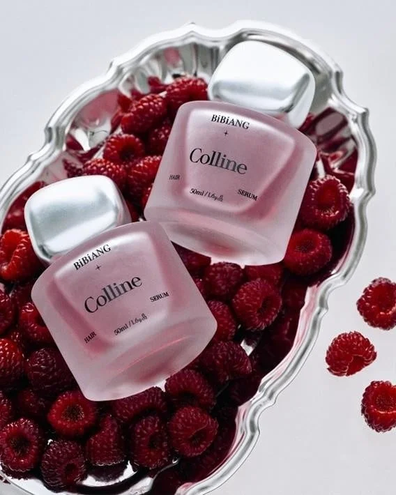 シルバーのトレイに山盛りの新鮮なラズベリーとともに、BIBIANG「Colline」のフロスト加工されたピンク色のヘアセラムボトルが2本美しく配置されています