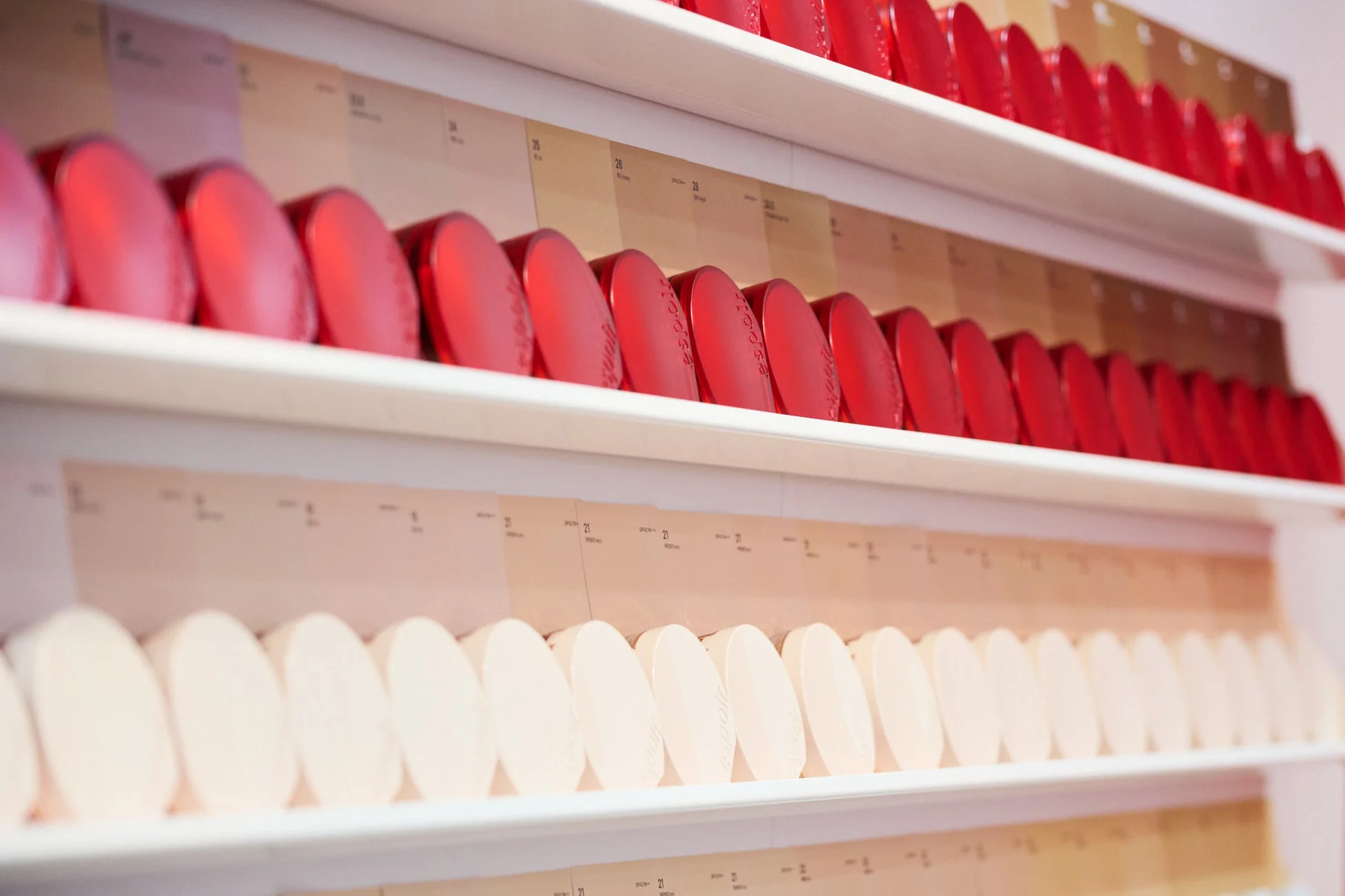 白い棚にESPOIRブランドの化粧品コンパクトが多数並べられたディスプレイ。赤やベージュ系の多様なシェードが色見本カードと共に展示されており、ファンデーションやパウダーの豊富なラインナップを示唆しています。