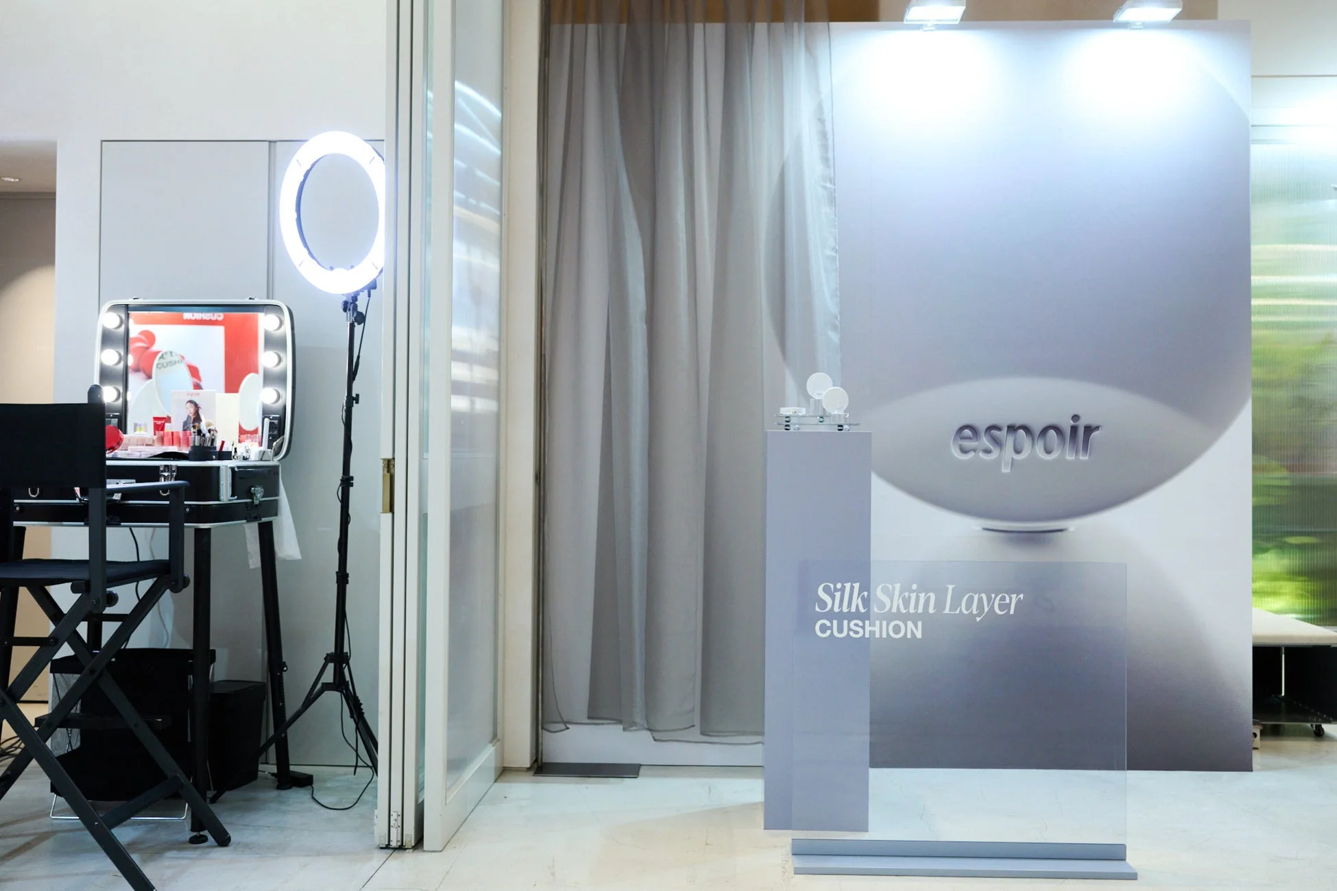 espoirブランドのコスメイベントまたはブースの様子を捉えた画像です。リングライトと電球付きのミラーがあるメイクアップステーションと、「espoir Silk Skin Layer CUSHION」のロゴが入ったディスプレイエリアが設置されています。