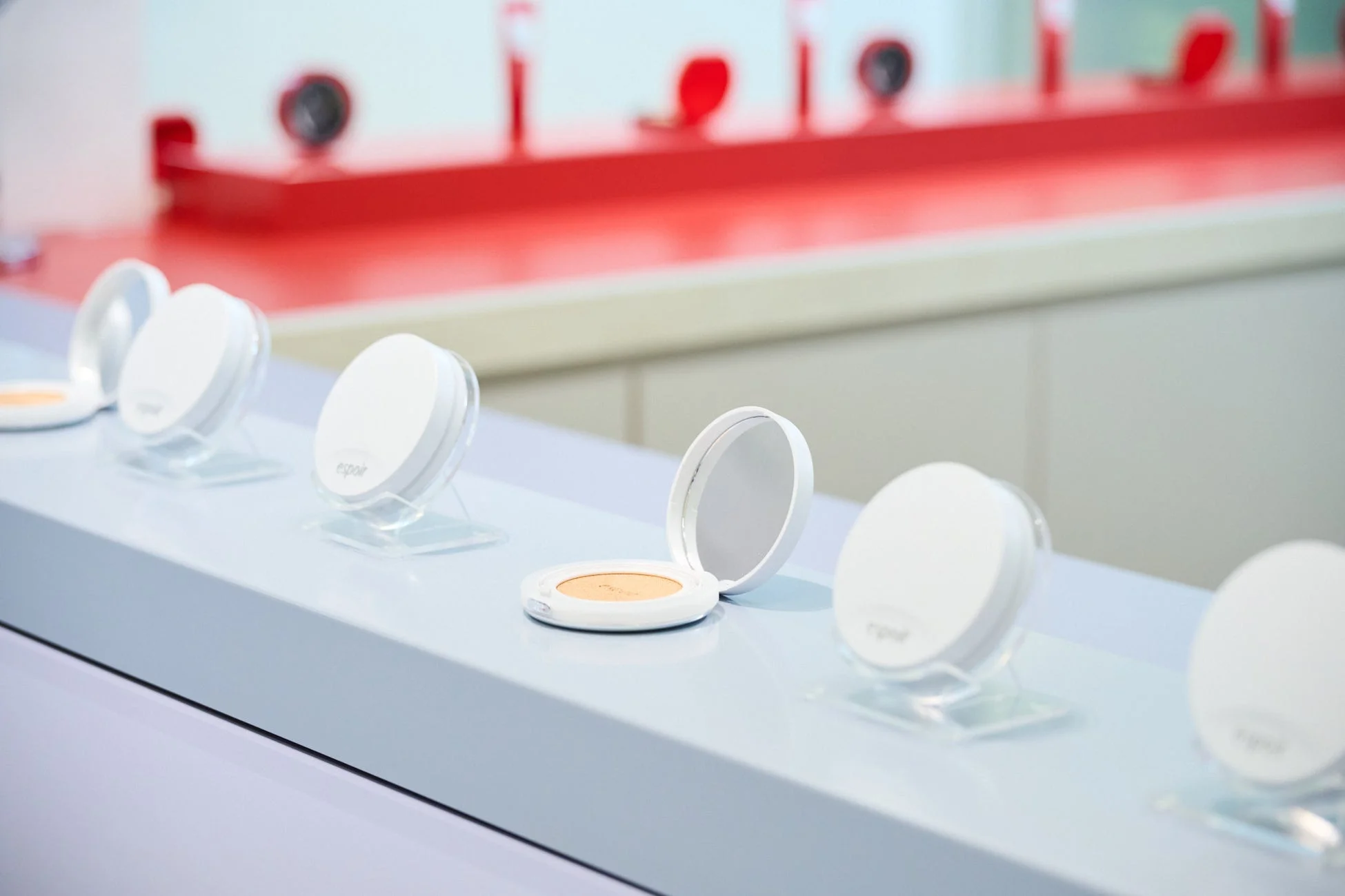 白いコンパクト型化粧品が複数、ライトブルーの陳列棚に並べられています。中央の1つは蓋が開いており、中身のファンデーションのような製品と鏡が見えます。