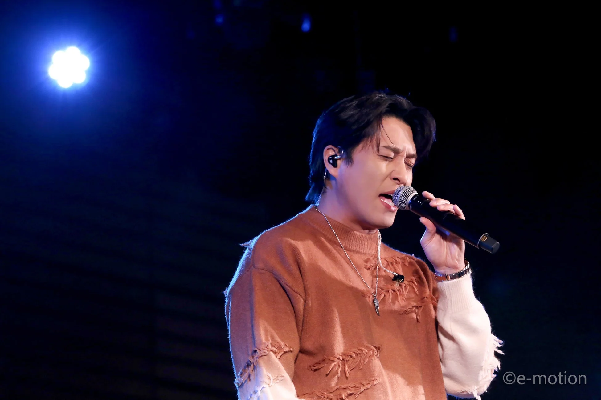 青いスポットライトが当たるステージで、男性歌手が目を閉じてマイクを握り熱唱している様子です。