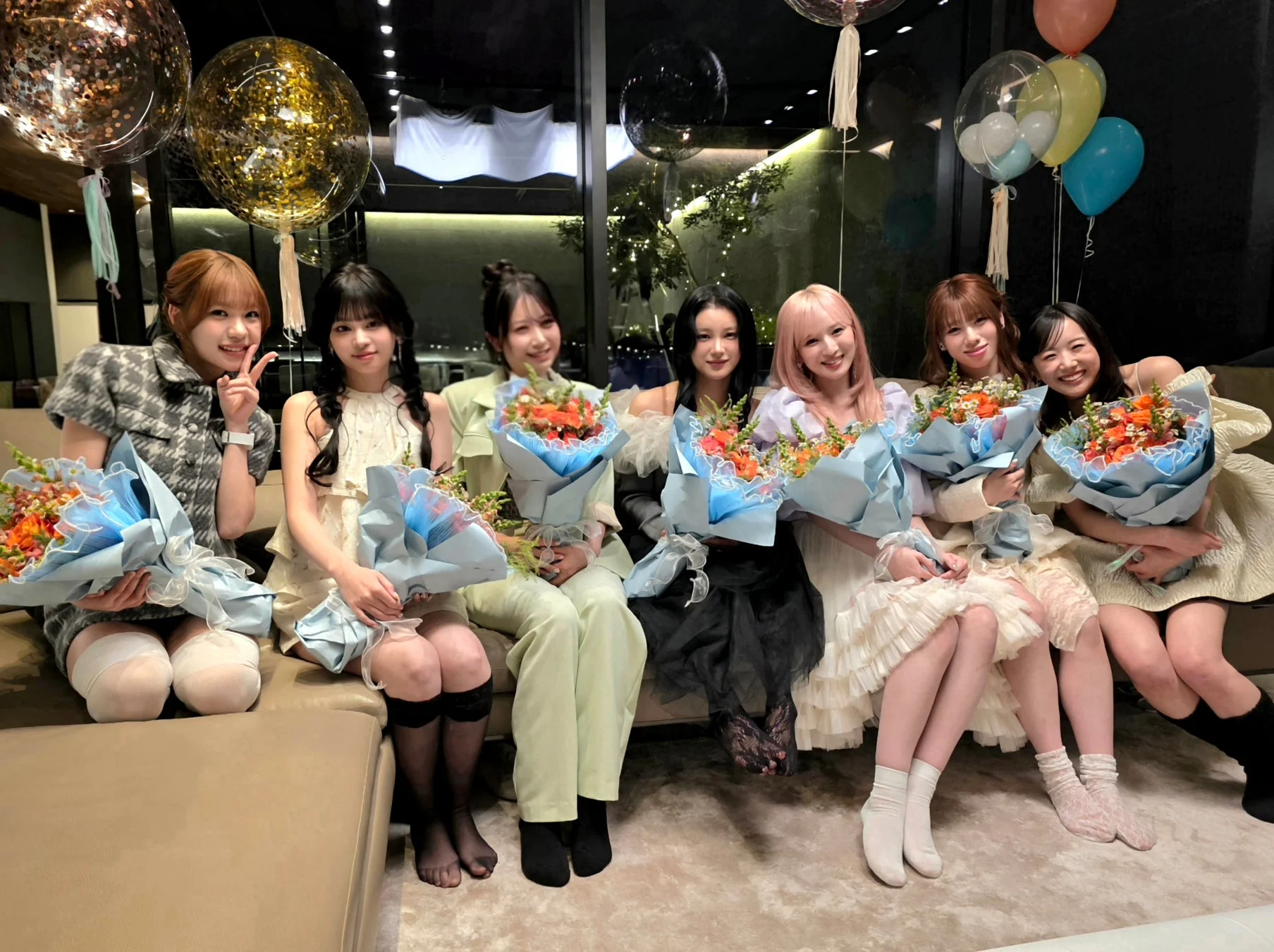 7人の女性が花束を持ち、笑顔でカメラにポーズをとっている集合写真です。背景にはバルーンが飾られており、お祝いやパーティーのような華やかな雰囲気が伝わります。