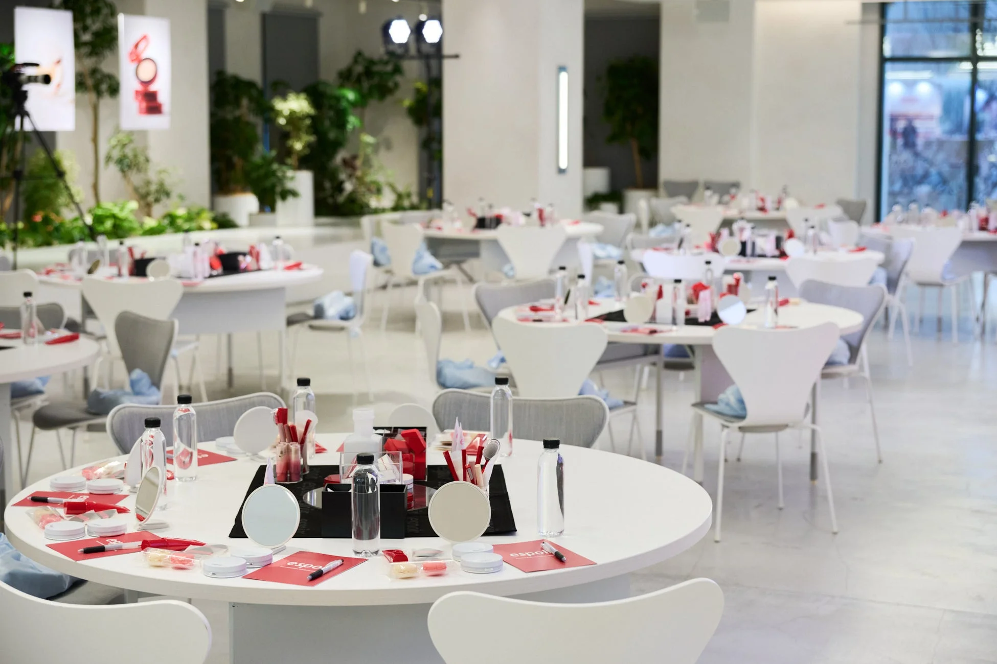 明るいモダンなイベント会場に、メイクアップワークショップ用の丸テーブルが多数並んでいます。各テーブルには化粧品、鏡、水ボトル、赤い用紙が用意され、背景には植物が見えます。