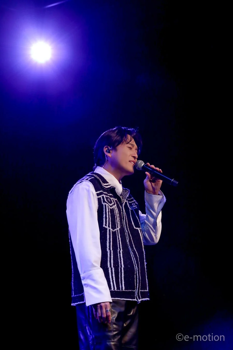 青いスポットライトを浴びた男性歌手が、ステージ上でマイクを手に熱唱している様子。