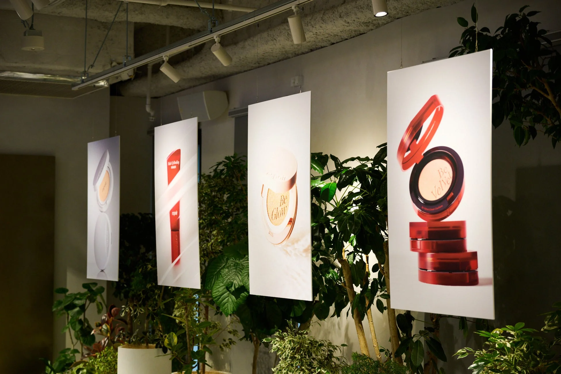 屋内空間に吊るされた複数のエスポア化粧品ポスターが展示されており、周囲には多くの植物が配置されています。Be GlowやBe Velvetなどの製品が紹介されています。