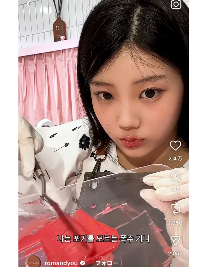 若い女性が白い手袋をはめ、真剣な表情で赤いリップカラーのようなものを透明なパレットで混ぜています。メイクアップ作業中の様子を捉えた画像です。