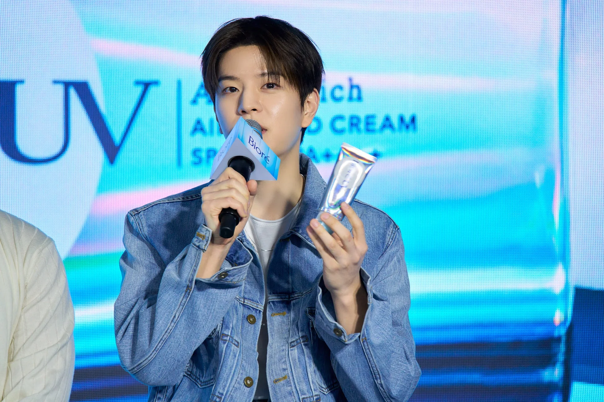 男性がステージ上でマイクを持ち、日焼け止めと思われる製品を手にしています。背景のスクリーンには「UV」「CREAM」「PA++」などの文字が見え、ビオレ製品のプロモーションイベントの様子です。