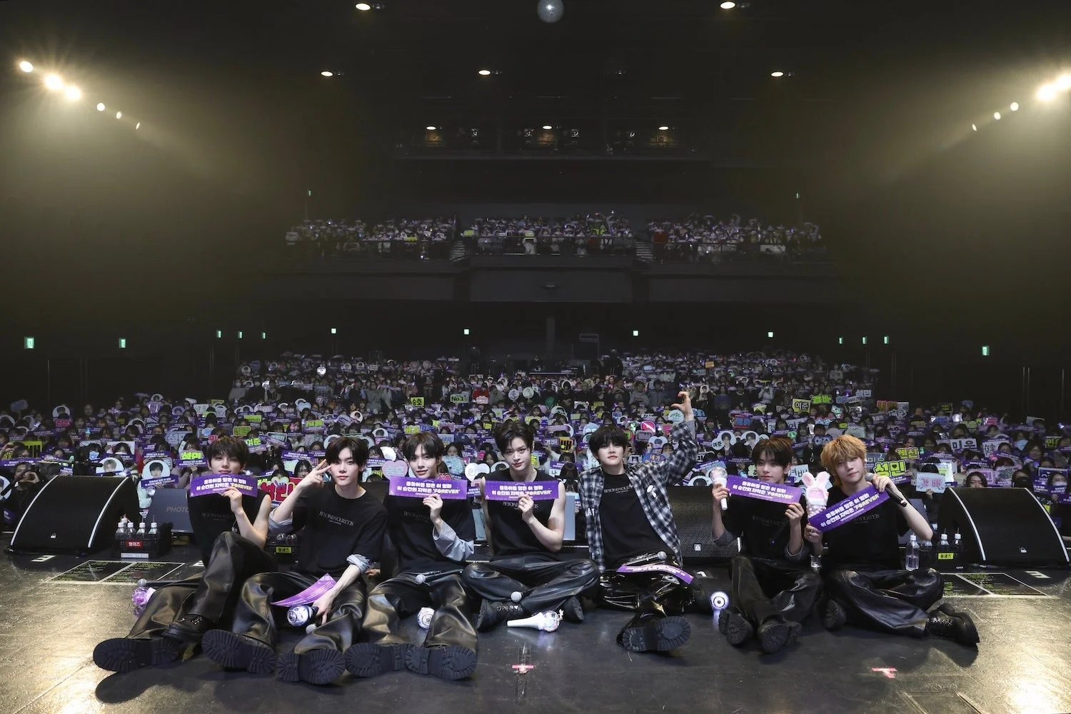 暗い会場のステージに座る男性アイドルグループが、大勢のファンを前に紫色のバナーを掲げている