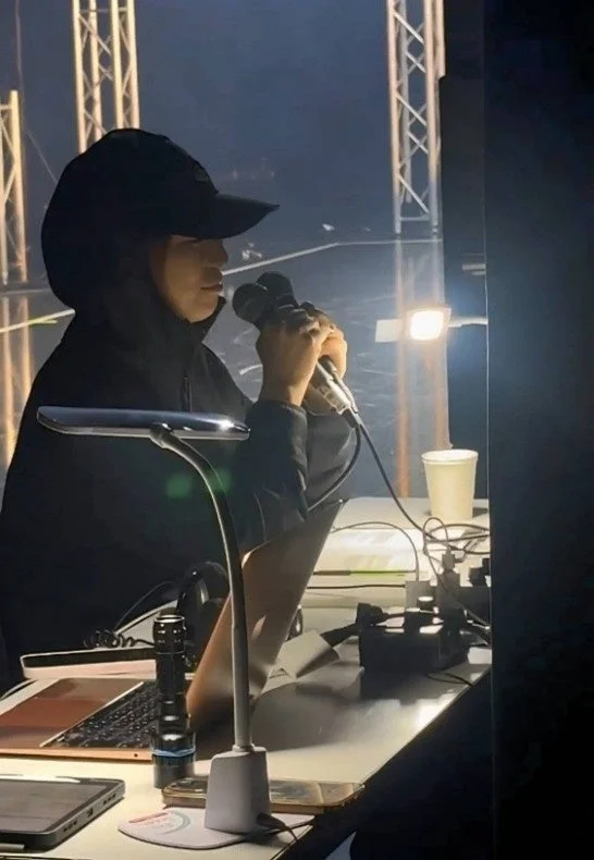 黒いパーカーとキャップを被った人物が、暗いステージ裏のような場所で2本のマイクを手にしている。デスクにはランプや機材が置かれており、イベントの音響スタッフかパフォーマーの準備風景と見られる。