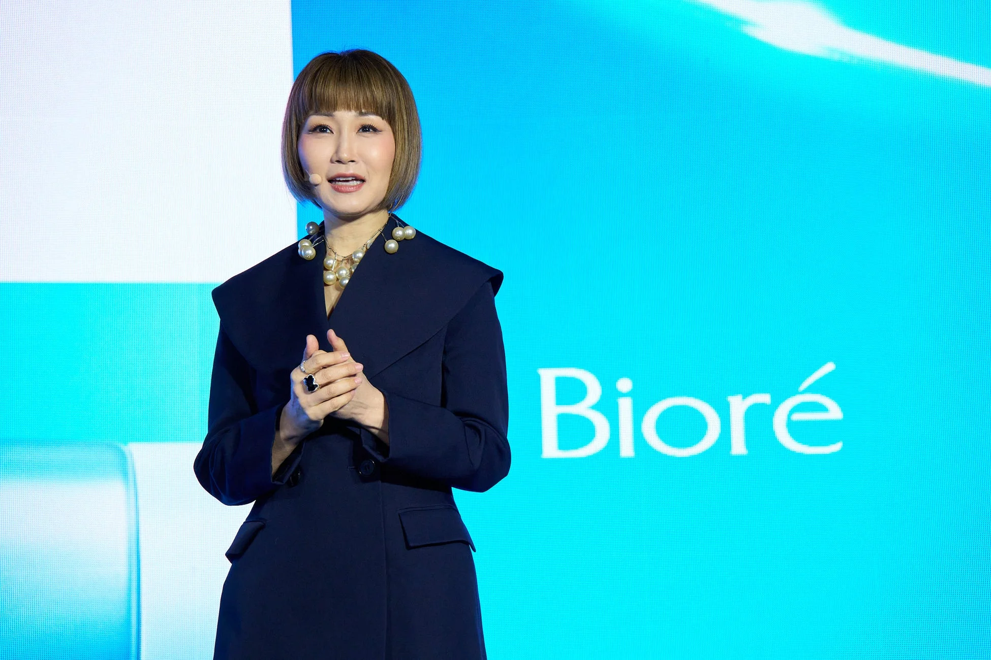 ステージ上で講演する女性の姿です。青い背景には「Bioré」のロゴが表示されており、女性はネイビーのコートとパールネックレスを身につけています。