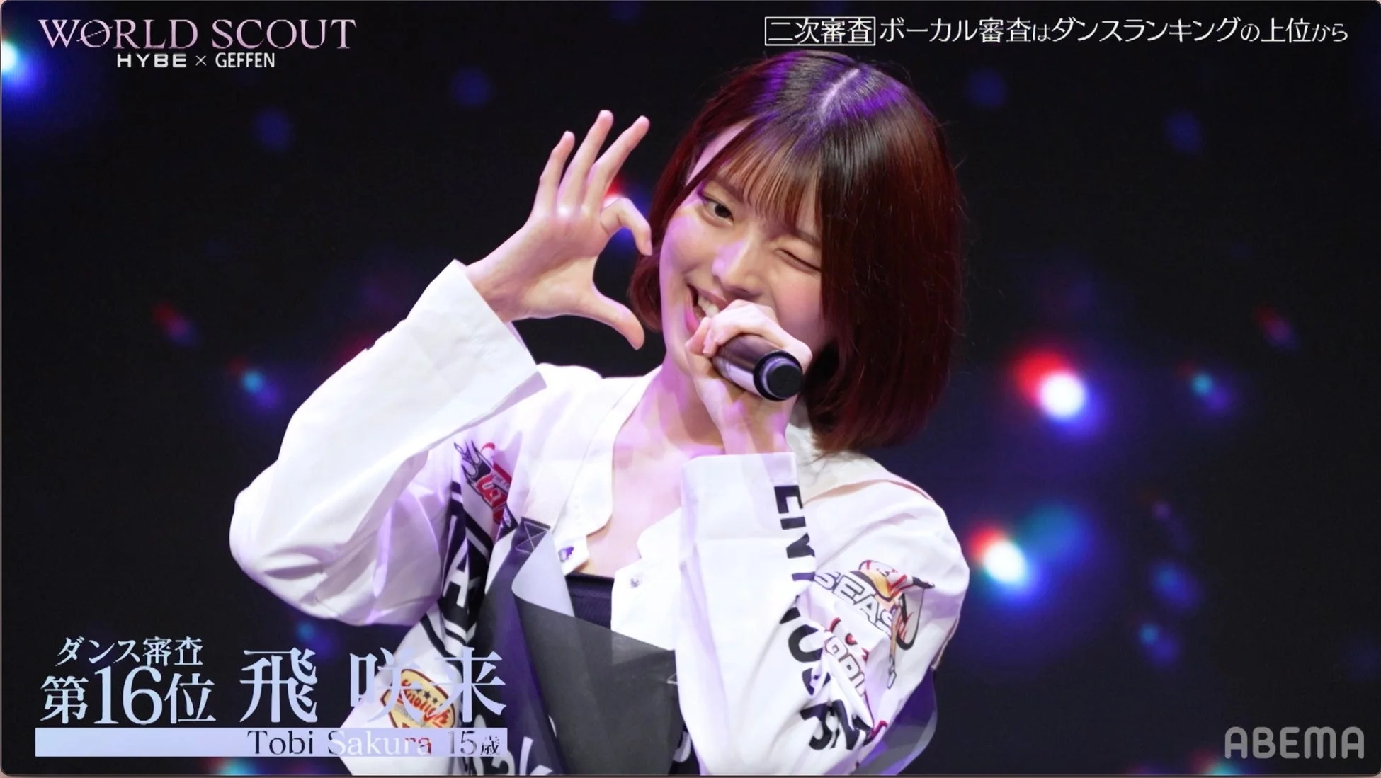 ステージ上でマイクを持ち、笑顔でウィンクしながらハートのポーズをする若い女性