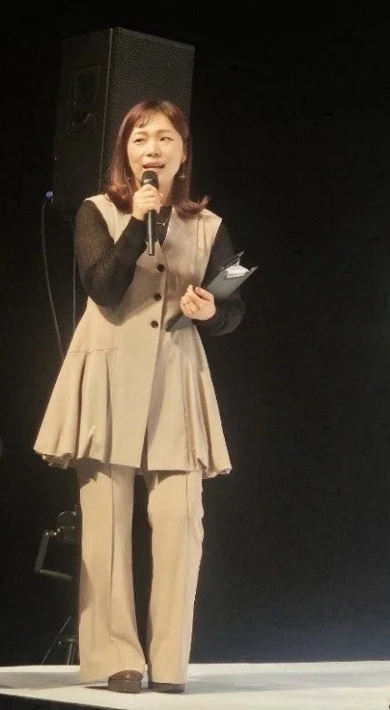 壇上でマイクを手にスピーチをする女性の姿。ベージュのベストとパンツを着用し、手には資料らしきものを持ち、聴衆に向かって話している様子がうかがえます。