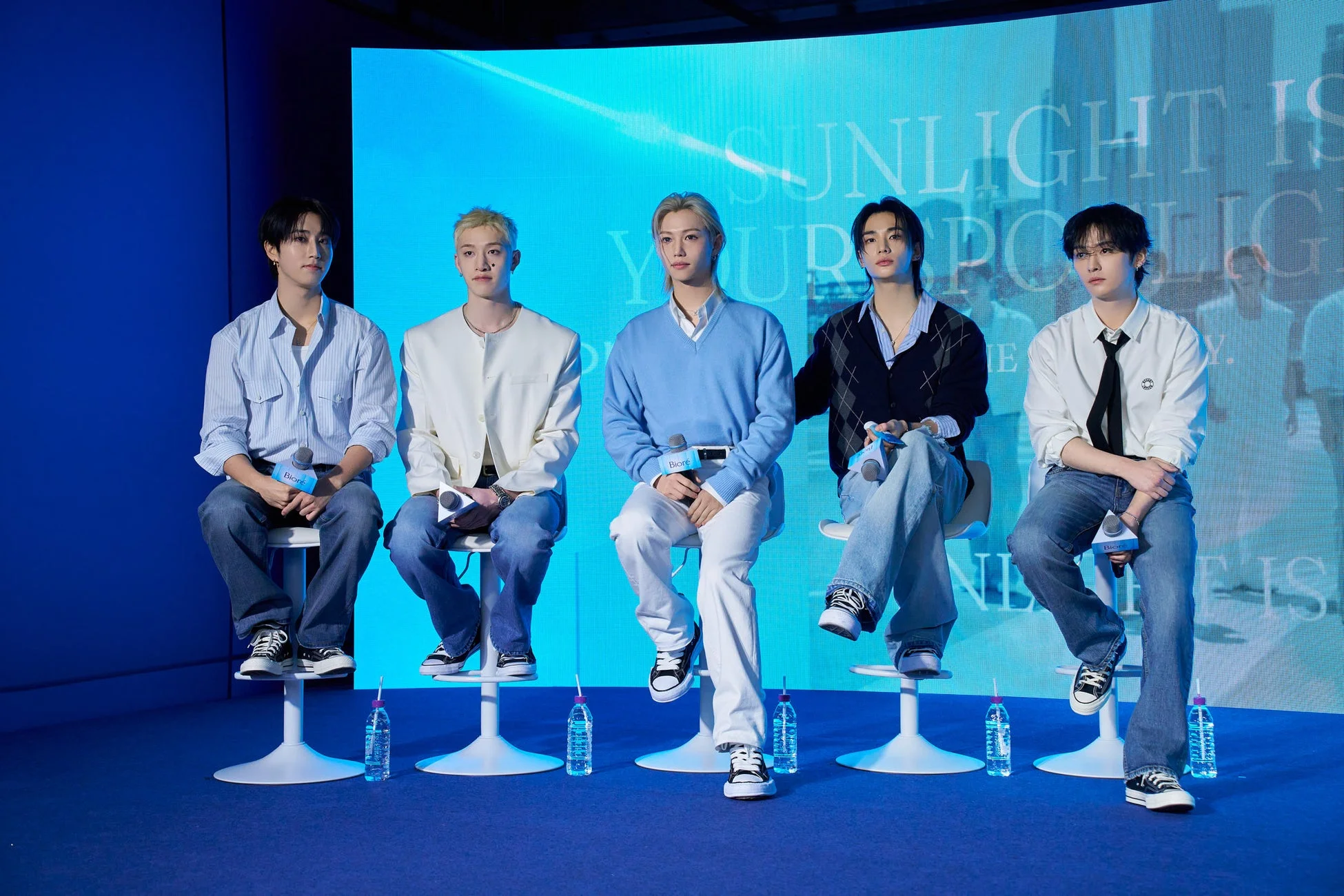 青い背景のステージで、男性アイドルグループのメンバーらしき5人が椅子に座り、マイクを持ってイベントに参加している様子。彼らはカジュアルな服装をしており、背景には「SUNLIGHT IS YOUR SPOTLIGHT」の文字が見える。