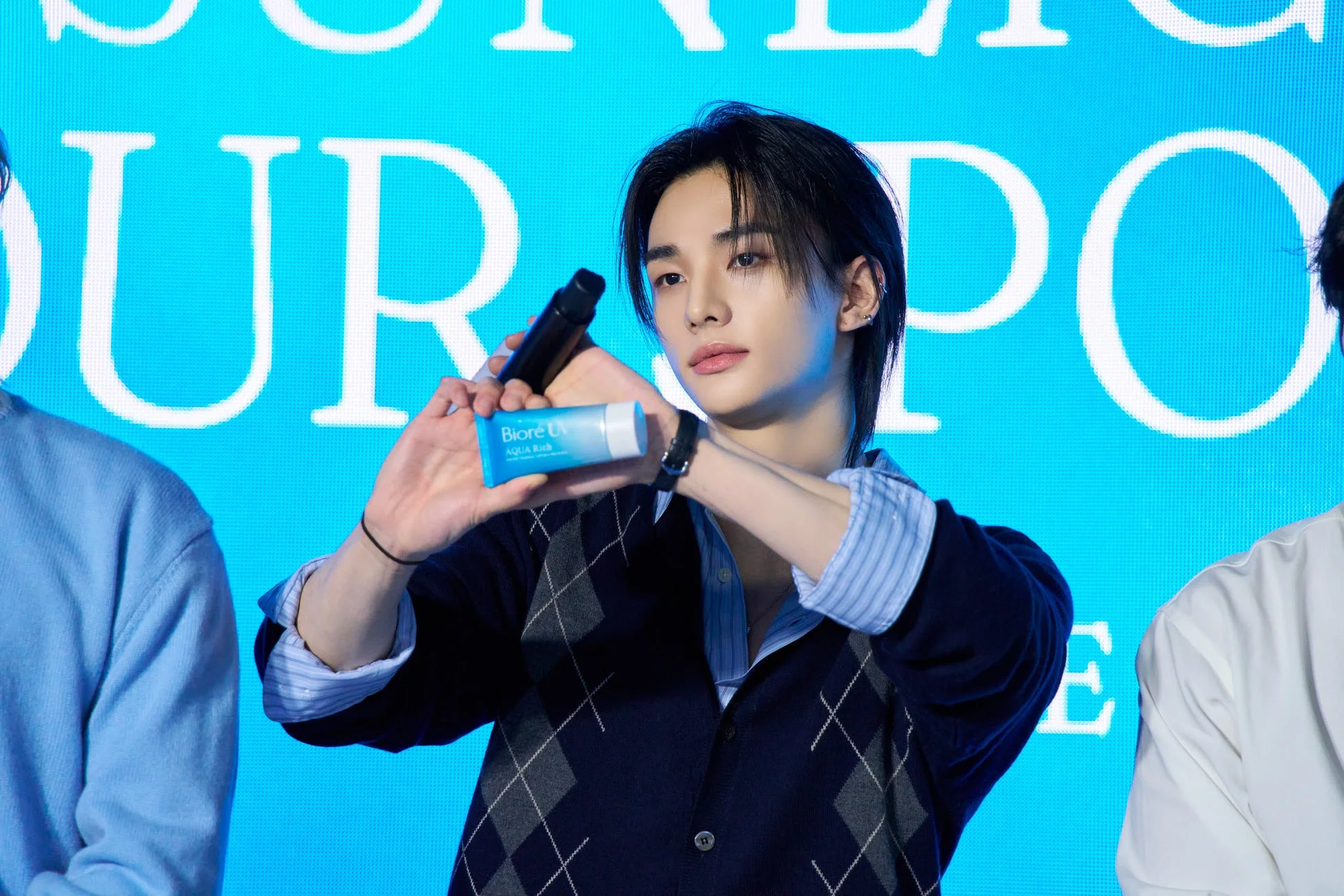 K-POPアイドルのような若い男性が、青い背景の前で「Biore UV AQUA Rich」の日焼け止め製品を手に持っています。彼はアーガイル柄のカーディガンを着ており、製品をアピールしている様子です。